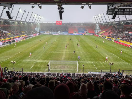 Pohled na probíhající fotbalový zápas z tribuny vlajkonošů za bránou. Fanoušci jsou na místech, ale nefandí.