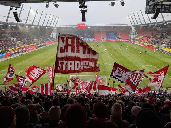 Pohled na probíhající fotbalový zápas z tribuny vlajkonošů za bránou. Fanoušci jsou na místech a intenzivně fandí, červenobílé vlajky vlají.