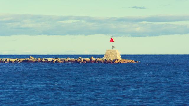A short obelisk with a triangular sign and probably a light is on the edge of a thine jetty of light colored boulders just below the horizon of a slightly choppy sea with languid clouds stretched horizontally above
