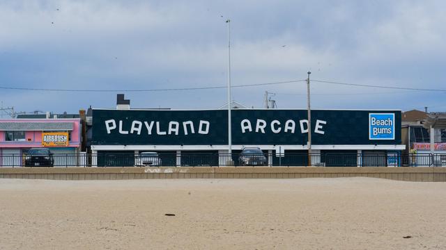 At the bottom is an almost clean sandy beach, then a concrete wall and a large green building with haphazard letters: PLAYLAND ARCADE in white with a blue sign on the upper right corner that says BEACH BUMS with a little purple shop to its right and a flagless flagpole in the exact center