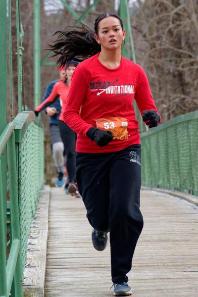 Woman wearing a red shirt and black pants is running with her black hair in a ponytail splayed to the the left with bib number 53 and black gloves.  She's running on a bridge with a floor of fine wooden horizontal slats with green fence sections and suspension cables above