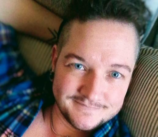 A trans man with dark hair and facial hair. He has piercing blue eyes, and a slight smile as he looks at you. He reclines on grey and white stripey pillows, and wears an unbuttoned blue checked shirt.