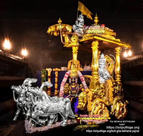 Idol of Hindu deity Krishna in bright purple seated in the driver seat of a horse-drawn gilded chariot with a silver figure of Arjuna in the passenger seat. The chariot is drawn by four silver horses [screen left] and has an ornate roof with a small umbrella at its front. At the leading edge of the roof, behind the umbrella, sits a silver Lord Hanuman holding up a gilded flag. A string of white flowers (some red bands) around Krishna. Arjuna sits with hands folded in a prayer pose and has a silver bow with him. Two rails of oil lamps on either side of the chariot.