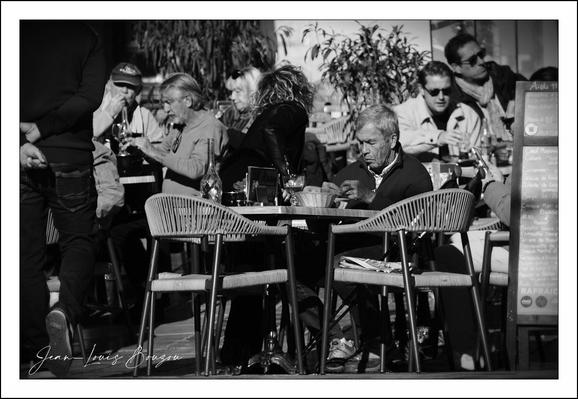 A black-and-white street photograph of a busy outdoor café, full of small round tables and woven chairs. In the foreground a man sits alone at a table, hunched slightly over a cup, his face lined and thoughtful as if lost in his own world. Around him the terrace hums with life: couples and small groups lean in to talk, a woman lifts a glass, another person gestures with a cigarette, sunglasses catching the light. Dappled sunlight and deep shadows give the scene strong contrast, bringing texture to coats, wicker seats and the leafy plants behind the diners. To the right a chalkboard or menu stands by the tables, anchoring the café setting; a photographer’s signature is visible in the lower-left corner. The overall mood is cinematic — a quiet, reflective moment set against the warm bustle of everyday city life.