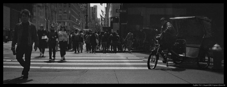 A rickshaw waits for passengers on the side of a curb with its driver seated on its bicycle as a crowd of people walk by through a nearby street crossing.