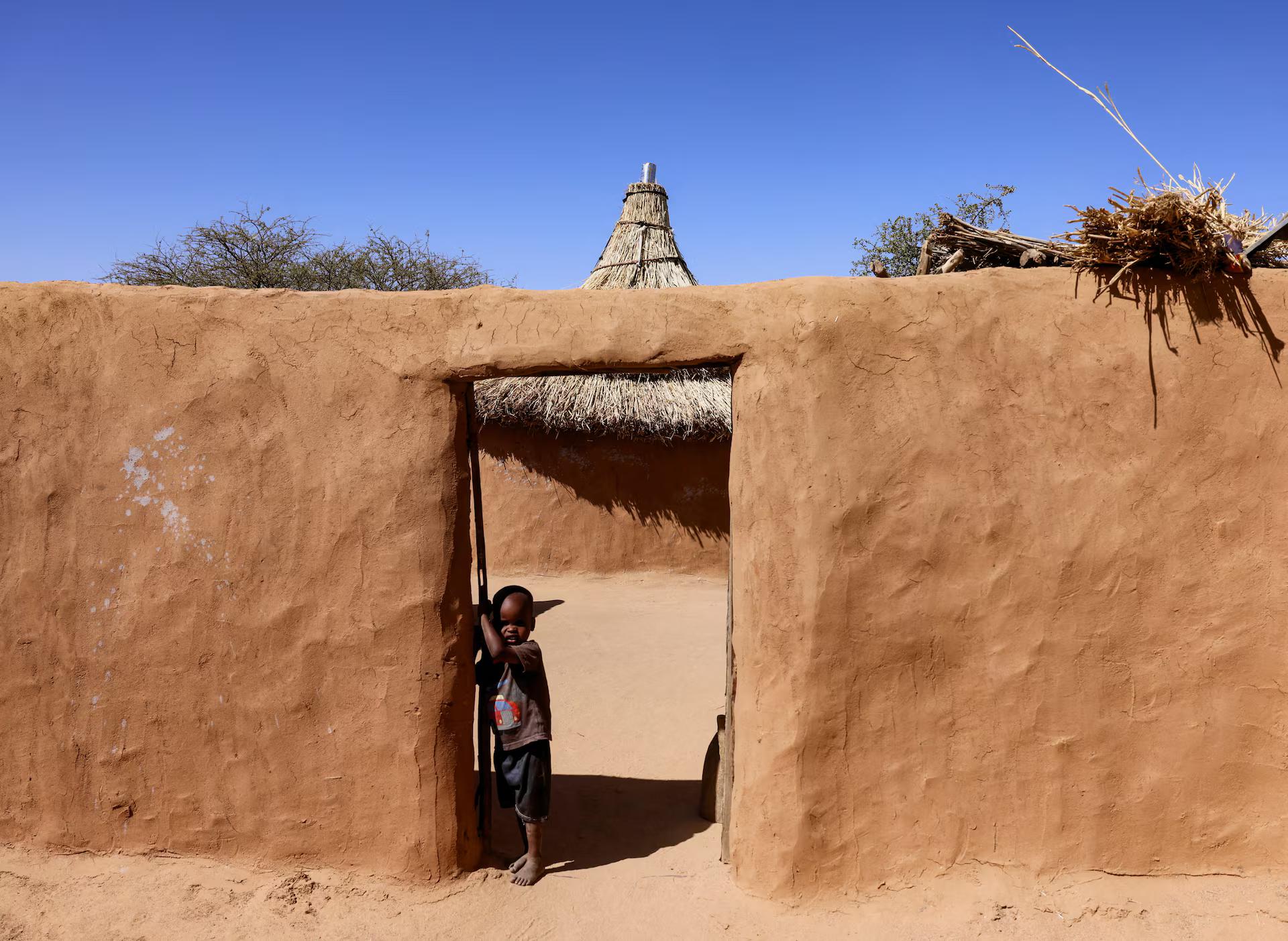 A Sudanese refugee boy from Darfur stands in front of his family’s mud house.