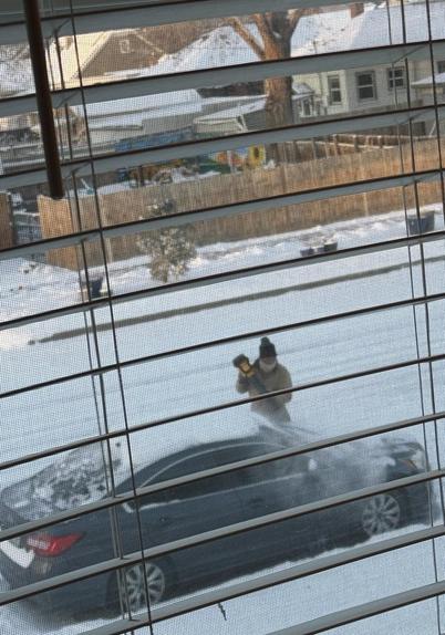 A man in a snow suit is using a small leaf blower to clear about 6" of snow off a gray car on a deeply snow covered street. The photo is taken from above  thru a window with the blinds down but turned open. Snow is seen on the houses across the street and for blocks beyond. 