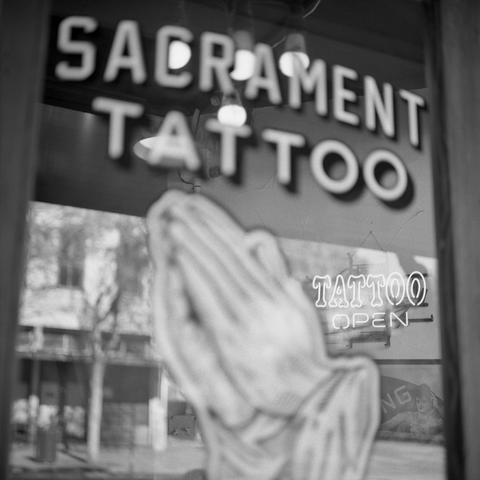 A black-and-white film photo. A neon sign, TATTOO OPEN, shines through the front glass door of Sacrament Tattoo in downtown Austin, Texas. Wednesday, November 26, 2025.