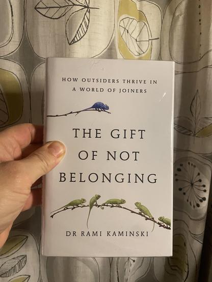 A white male hand holding a copy of 'The Gift of Not Belonging' by Rami Kaminski, in front of a curtain with a 1950s style pattern.