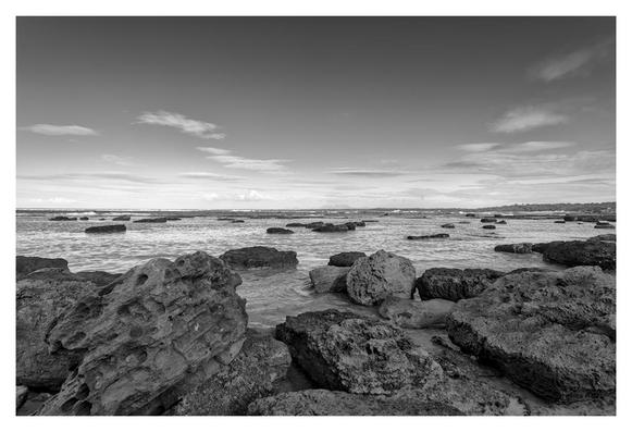 The black-and-white photograph shows a calm, expansive seascape. In the foreground, large, rugged volcanic rocks dominate the scene, some partially submerged in shallow, gently rippling water. The rocks are dark, rough-textured, and irregularly shaped, and scattered across the lower third of the image.Beyond the rocks, the ocean stretches out toward the horizon, its surface smooth and reflective with only subtle waves. Small clusters of rocks and low reef-like formations break the water’s surface here and there, creating a dotted pattern that leads the eye into the distance.The horizon line is low and perfectly straight, with a faint, distant shoreline barely visible under a vast, open sky. The sky takes up more than half the image and is filled with soft, wispy clouds. The overall mood is serene, quiet, and slightly moody due to the monochrome tones, and evokes a sense of peaceful isolation at the edge of the sea.