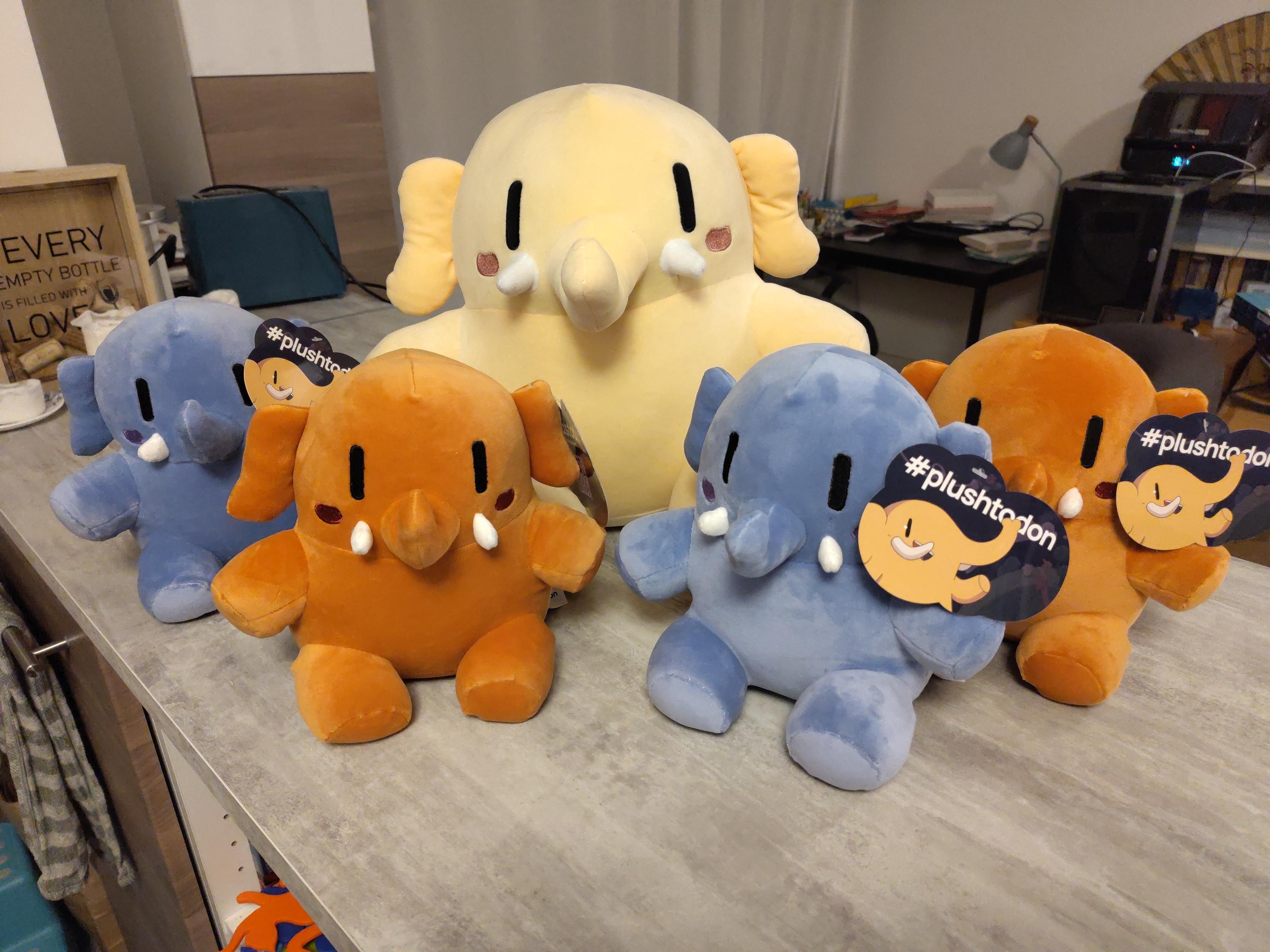 Cinq peluches Mastodon sur une table. Le gros jaune au milieu, et quatre petits autour, deux bleus et deux marrons.