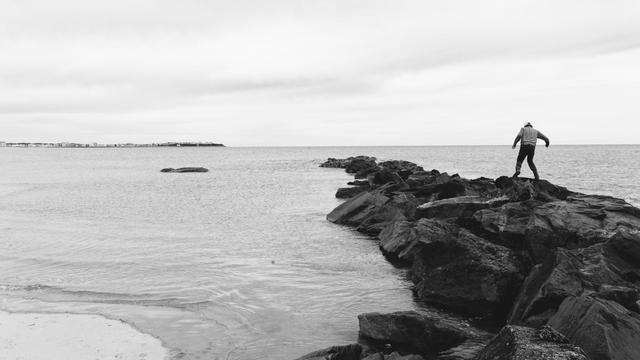 A man looks tiny in the context of a rock jetty he is walking across made of very dark rock in this black and white photo