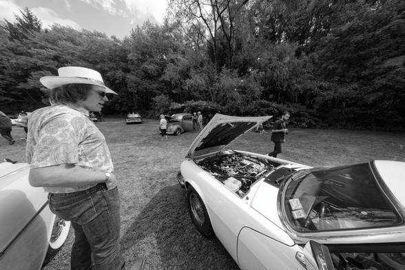 Man at the lower left with a light colored hat looks at a white car that has it's hood opened the wrong way.