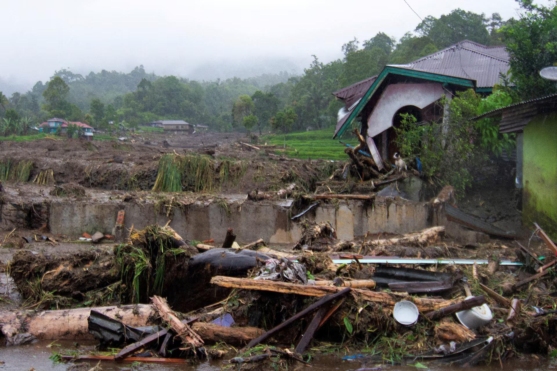Fallen tree branches and debris lie on the ground near a damaged house hit by flash floods.