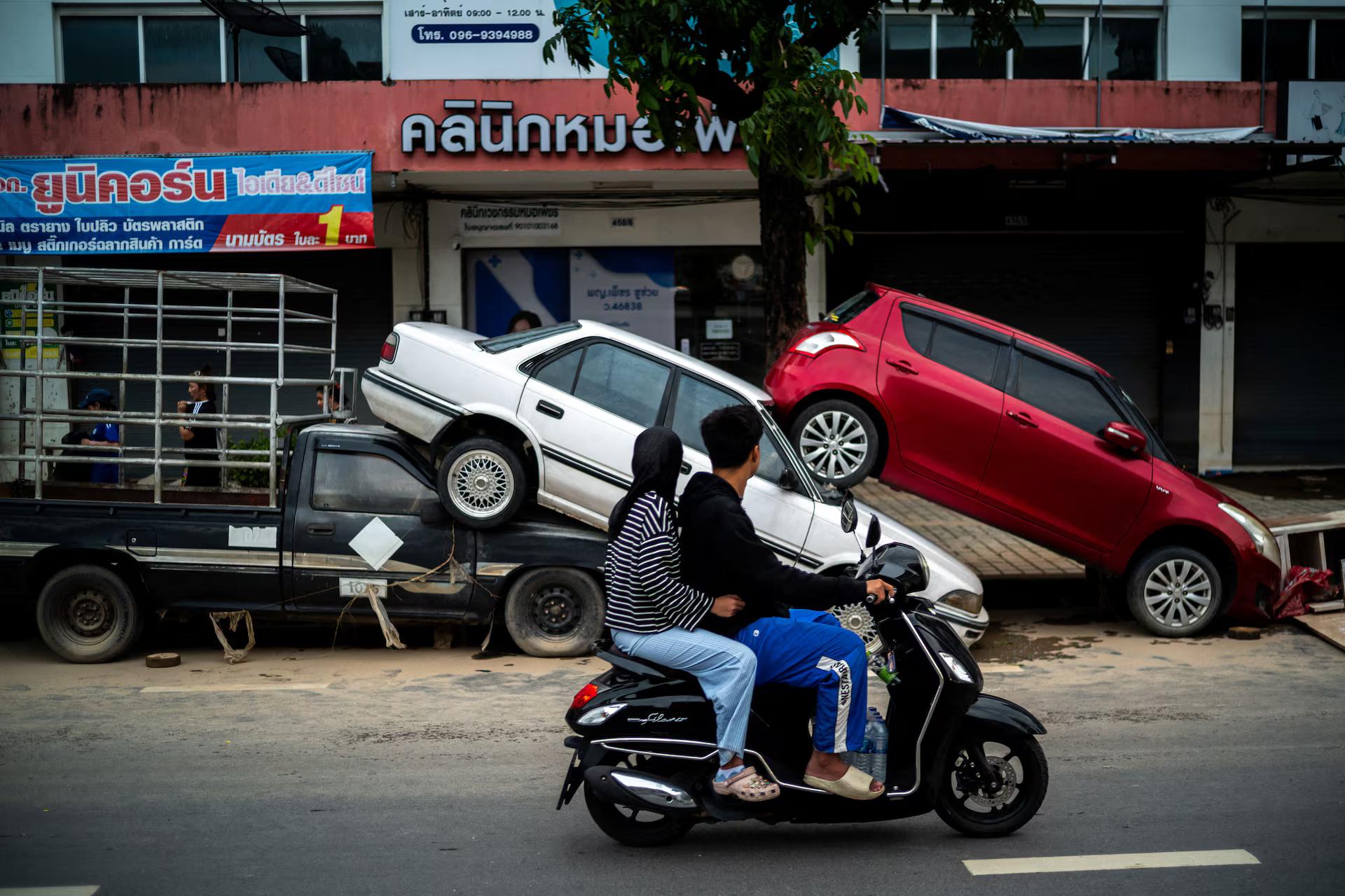 People ride a motorcycle past cars stacked up by the flood.