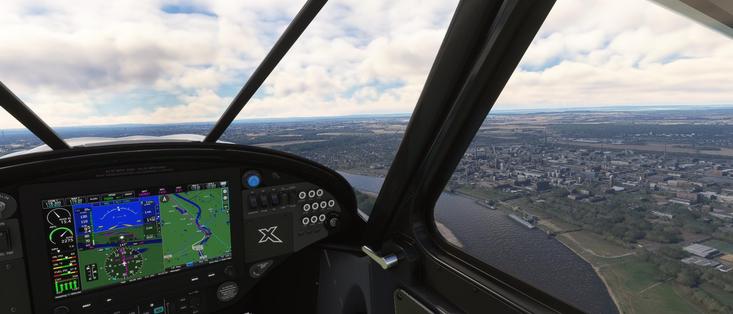 The image depicts a view from inside a cockpit of an aircraft, likely a small private plane, as seen from the perspective of the pilot or a passenger. The cockpit is equipped with modern avionics, including a digital display panel that shows various flight instruments and navigation data. The display includes information such as airspeed, altitude, heading, and other flight parameters. The outside view through the cockpit window shows a landscape with a river, fields, and a cityscape in the distance. The sky is partly cloudy, suggesting a clear day with some scattered clouds. The overall scene suggests that the aircraft is in flight, possibly over a rural or semi-urban area. The image appears to be from a flight simulation software, as indicated by the detailed and realistic graphics.