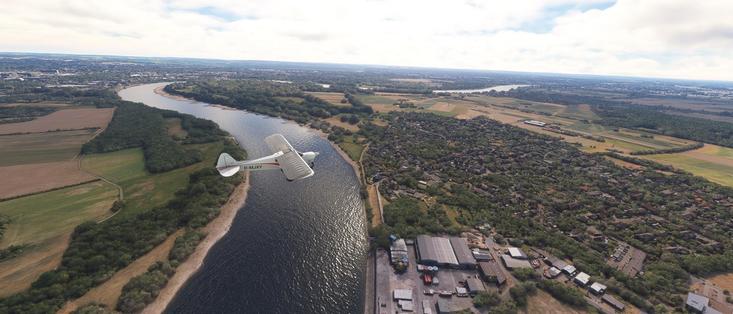 The image depicts an aerial view of a landscape featuring a small airplane flying over a river and a town. The airplane is a CubCrafters NXCub with the registration number "D-MJXY" visible on its side. The river curves through the landscape, bordered by green vegetation and fields. On the right side of the image, there is a town with buildings and industrial structures, including what appears to be a large warehouse or factory. The sky is partly cloudy, and the overall scene suggests a peaceful, rural setting with a mix of natural and developed areas. The image appears to be a screenshot from a flight simulation game, given the detailed rendering of the environment and the airplane.