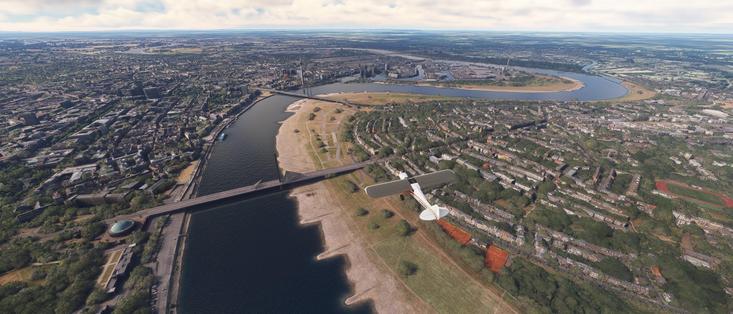 The image depicts an aerial view of a cityscape, likely captured from a flight simulation or a high-resolution satellite image. The scene includes a river running through the center of the image, with a bridge spanning across it. The river is surrounded by greenery and urban development, with buildings and roads visible on both sides. In the foreground, there is a small aircraft flying over the river, adding a dynamic element to the image. The sky is partly cloudy, and the overall scene is well-lit, suggesting it is daytime. The image provides a detailed view of the urban environment, including residential areas, commercial buildings, and natural landscapes.