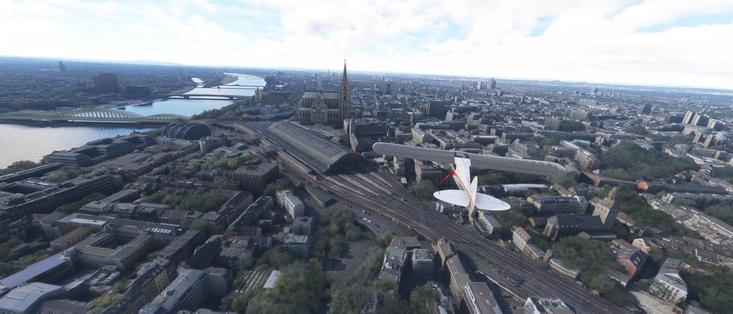 The image depicts an aerial view of a cityscape, likely taken from a simulation or a video game due to the high level of detail and the presence of a small aircraft in the foreground. The city features a prominent cathedral with a tall spire, which is a distinctive landmark. The cathedral is situated near a river, with bridges crossing over it. The surrounding area includes a mix of modern and older buildings, with some green spaces and trees interspersed throughout the urban environment. The sky is partly cloudy, suggesting a bright day. The overall scene is detailed and realistic, capturing the essence of a large, bustling city.