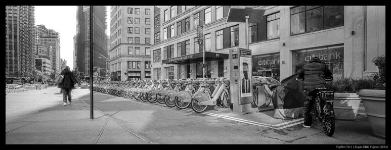 Divided by a street sign, two people take two different modes of transport through the city.  The person on the left walks away from the camera, while the person on the right is mounting a heavy bicycle for delivery.  Between the two are a row of docked Citibikes that stretch back into the distance.  The Flatiron building is visible in the background.