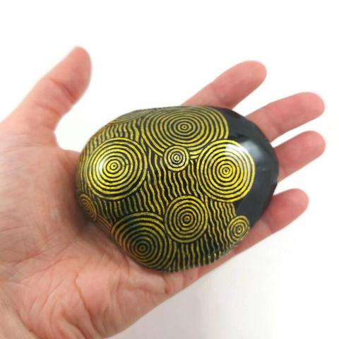 Galet noir recouvert de cercles concentriques et de lignes ondulées dorés.
Il est inspiré des motifs antiques présents dans le jeu vidéo "Zelda : Tears of the Kingdom". 
Il est tenu dans le creux d'une main ouverte.