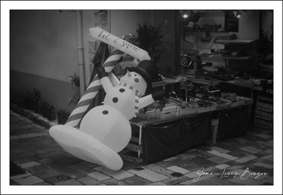 A black-and-white street photograph that feels quietly melancholy and a little whimsical. An oversized inflatable snowman — top hat perched, scarf around its neck and three big buttons down its front — leans awkwardly against a striped pole, as if it has just been toppled by a gust of wind. A small banner wrapped around the pole reads “Let it snow,” its cheerful message at odds with the snowman’s deflated, half-sitting posture. Behind them a shop window and a cluttered sidewalk display of goods provide a cluttered urban backdrop; a potted plant and crates add to the scene’s everyday detail. The contrast between the festive prop and the ordinary storefront, captured in grayscale, gives the image a quietly comic, slightly forlorn holiday mood. A photographer’s signature sits in the lower right corner.