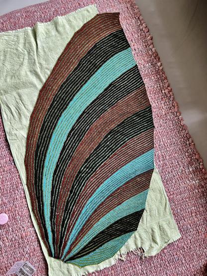 Drop or leaf shaped knitted fabric pinned out on a pink carpet with an old light green towel on it. The shawl is wider than the towel. It has stripes of green with three other colours. There are 12 segments. Green paired with turquoise, green paired with red brown and green paired with dark brown. So each combination appears four times.