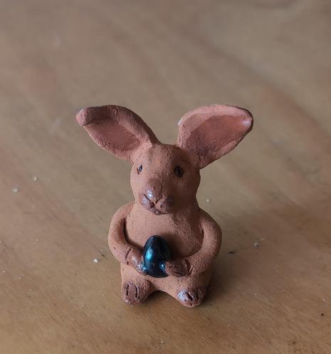 A little Christmas bunny made from clay sitting on a wooden desk. It has big ears and is, for some reason, holding an egg.