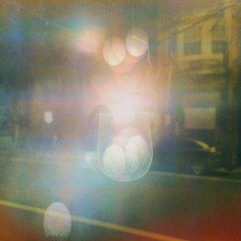 Objects that appear to be eggs appear in this streetscape which includes buildings, utility poles and wires, and a car. Some of the eggs are enclosed in a a glass light bulb and on it's wire. One stands in a little nest of vegetation on the curb. There is an eerie, dreamlike illumination. There is a surreal quality to the whole affair.