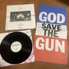 Das Bild zeigt eine auf der Seite liegende Schallplatte, ein quadratisches Albumcover mit einem Foto von Menschen und Fahrzeugen und ein großes Poster mit dem Schriftzug „GOD SAVE THE GUN” in fetten, bunten Buchstaben.