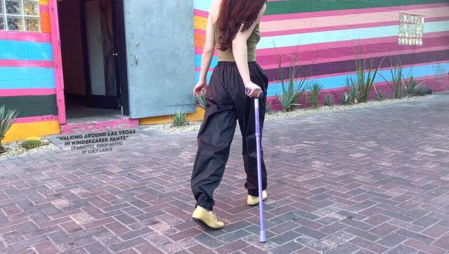 A still from a video by Lucy LaRue showing the petite brunette with long curly hair using a purple cane to walk down a brick path along a colorful striped painted wall of varying purples and blue hues with a desert plant edging, wearing black wind breaker pants, a green tank top, and tan shoes.  The credits "Walking Around Las Vegas In Windbreaker Pants" (2 minutes, 1080p 60fps) are written on the image underneath an open doorway which clearly beacons to the young woman as she walks.