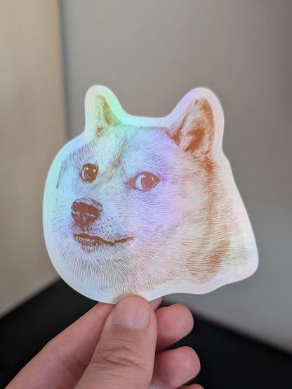 Photo of a left hand holding a diecut sticker. The sticker is a holographic drawing of the Shiba named Doge/Kabosu.
