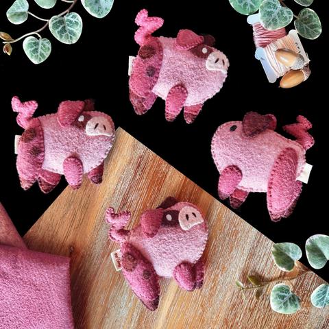 Four little handmade felt pigs on a black and wooden background with decorative plants. Every piggy is the same. Their bodies are round and light pink, they are looking up with pale pink snouts with embroidered nostrils, a brown mud spot around their white eyes. Their ears are droopy, pink on the front side, a shade darker than the body’s and the same as the legs and tail. The back ears are muddy brown. The legs have little brown hooves and two little mud spots on its thigh.