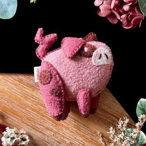 A little handmade felt pig photographed on a wooden and black bag round, surrounded with plants and flowers on each corner: its body is round and light pink, it’s looking up with a pale pink snout with embroidered nostrils, a brown mud spot around its white eye. It’s ears are droopy, pink on this side, a shade darker than the body’s and the same as the legs and tail. The legs have little brown hooves and two little mud spots on its thigh.