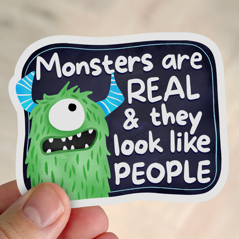 A sticker showing a cute green one-eyed cartoon monster with blue striped horns next to the text ‘Monsters Are Real & They Look Like People.’ A hand is holding the sticker.