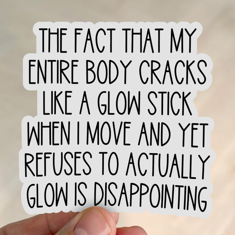 A hand holds a white sticker with black playful lettering that reads, “The fact that my entire body cracks like a glow stick when I move and yet refuses to actually glow is disappointing.”