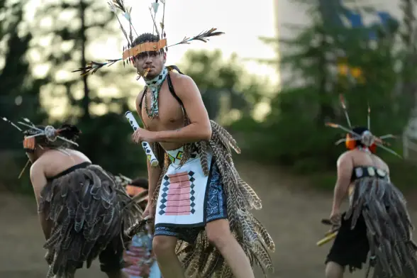 A dancer wearing feathered regalia and an orange-and-white headdress performs outdoors, holding ceremonial items, with other similarly dressed dancers moving in the background.