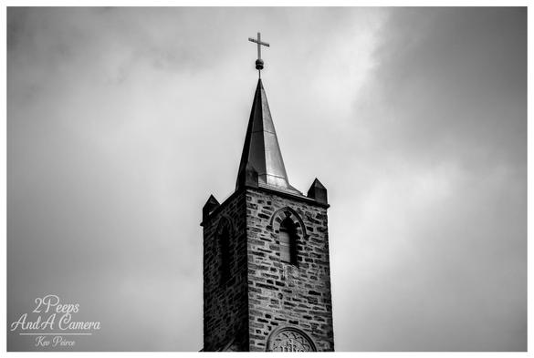 Black and white photograph showing the tall, stone steeple of a church in Burra, South Australia, topped with a cross.
The spire is dramatic against a cloudy, atmospheric sky, emphasizing the architectural details of the bell tower.