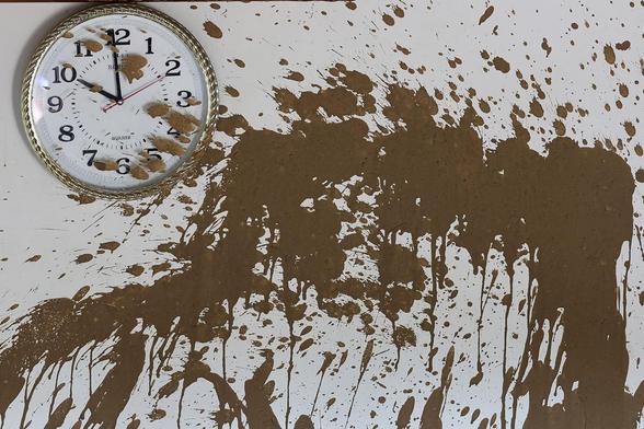 A clock hangs on a wall covered with mud inside a house.