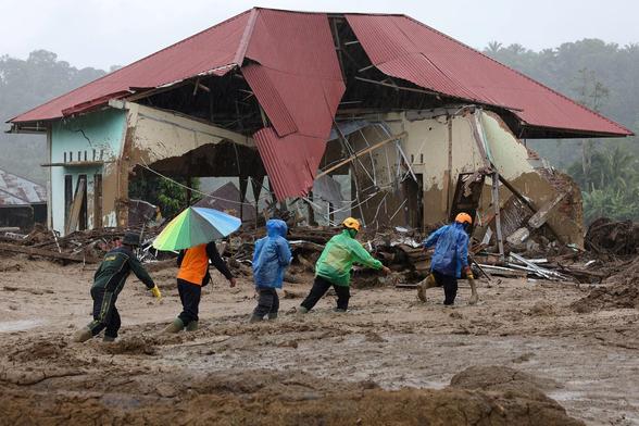 Rescuers walk through the mud past a house damaged by flood.