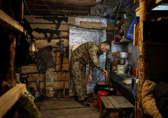 A serviceman prepares food in a dugout, with guns hanging on the wall.
