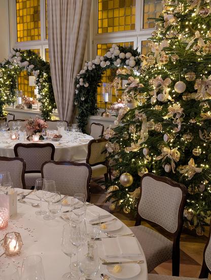 An elegant dining room set for a formal dinner. Round tables are covered in white linens with wine glasses, silverware, and glowing votive candles. To the right, a large Christmas tree is decorated with gold ornaments and cream-colored ribbons. in the background, tall windows are framed with lush green garlands and white baubles.