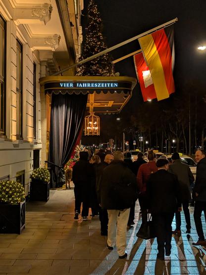 The exterior entrance of the "Vier Jahreszeiten" hotel at night. A crowd of people is gathered near the entrance under a black awning featuring the hotel's name in illuminated letters. A large German flag and a Hamburg state flag hang prominently from the building facade, illuminated against the dark sky, while a lit Christmas tree is visible on the balcony above.