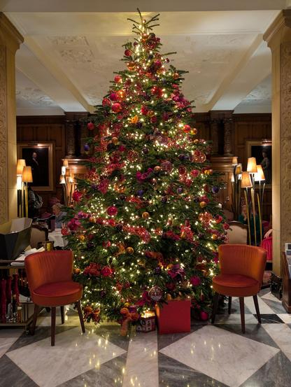 A tall, lavishly decorated Christmas tree stands as the centerpiece of an elegant room with marble floors and wood-paneled walls. The tree is glowing with warm fairy lights and adorned with red, purple, and gold ornaments. Two plush, burnt-orange velvet chairs sit symmetrically on either side of the tree, and red gift boxes are placed at the base.