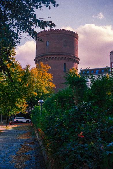A tall, cylindrical red brick water tower with battlements, set against a cloudy pink and blue sky, framed by trees in autumn foliage, with a paved road and a car visible in the foreground.
