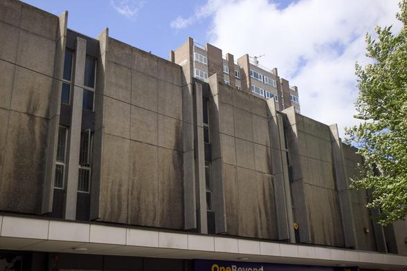 A Brutalist concrete building with tall, narrow vertical elements and dark stains beneath the windows. A modern block of flats is visible in the background, and a leafy tree is on the right under a partly cloudy blue sky.