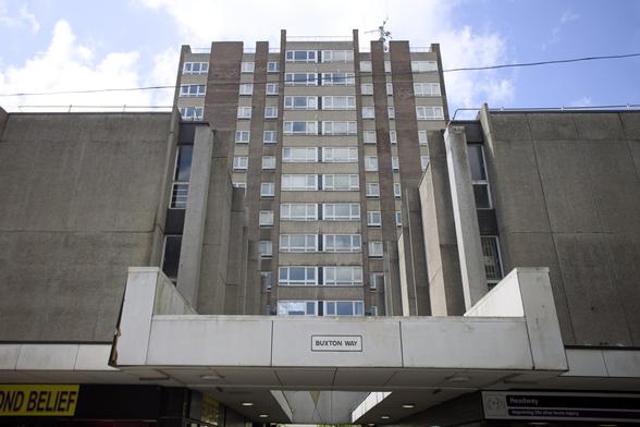 A tall, grey concrete block of flats with many windows stands behind a modern entrance labelled Buxton Way. The structure has a Brutalist architectural style, with boxy shapes and minimal decoration, beneath a partly cloudy blue sky.