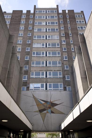 A tall, brown and grey block of flats with many rectangular windows rises into a blue sky. At the base, a geometric mosaic art piece decorates the wall, featuring intersecting triangles in shades of blue, yellow, brown, and cream.