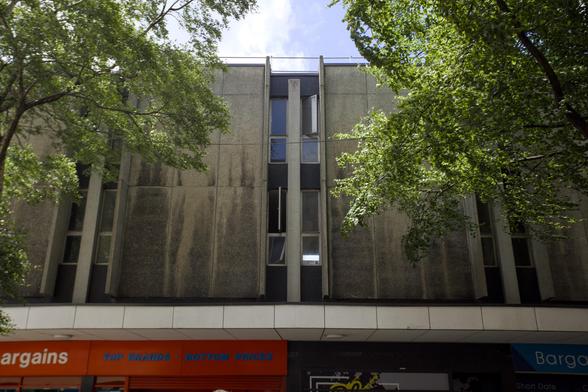 A grey, weathered concrete building with narrow vertical windows is partially shaded by leafy trees. At street level, there are red and blue shop signs, including one reading Bargains. The sky above is bright and partly cloudy.