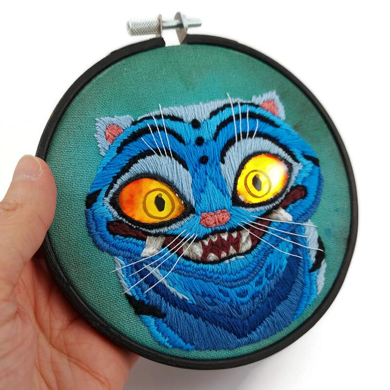 Broderie de Derpy, le tigre bleu démoniaque du film "Kpop Demon Hunters".
Elle est réalisée sur une toile verte foncée, tendue sur un cercle à broder en bois noir.
Les yeux sont des morceaux de CD fixés à la toile avec la technique de la broderie miroir, puis recouverts de résine teintée dans un dégradé jaune-orange-rouge.